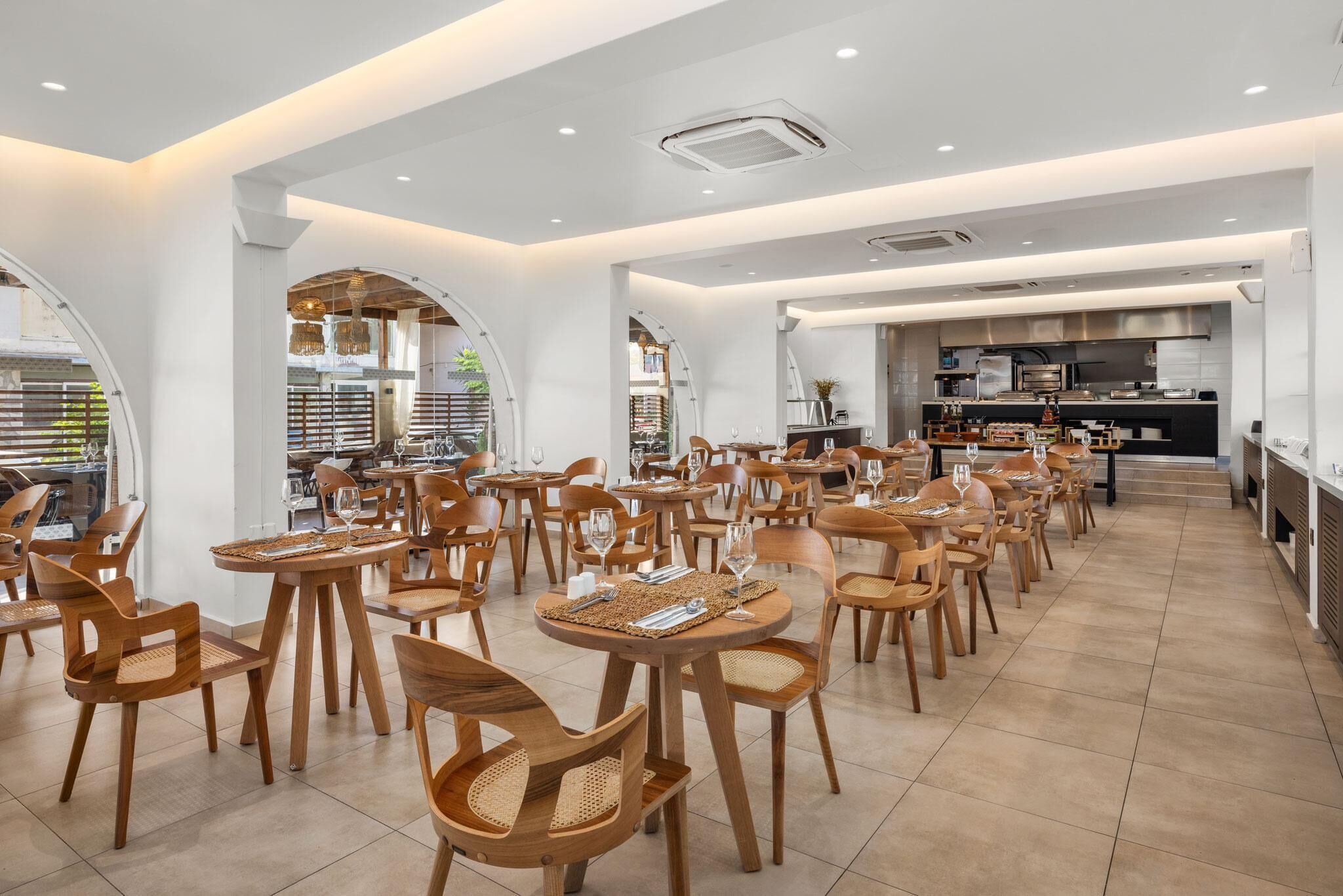A spacious dining area with wooden chairs and tables, a kitchen in the background, and a modern interior design.
