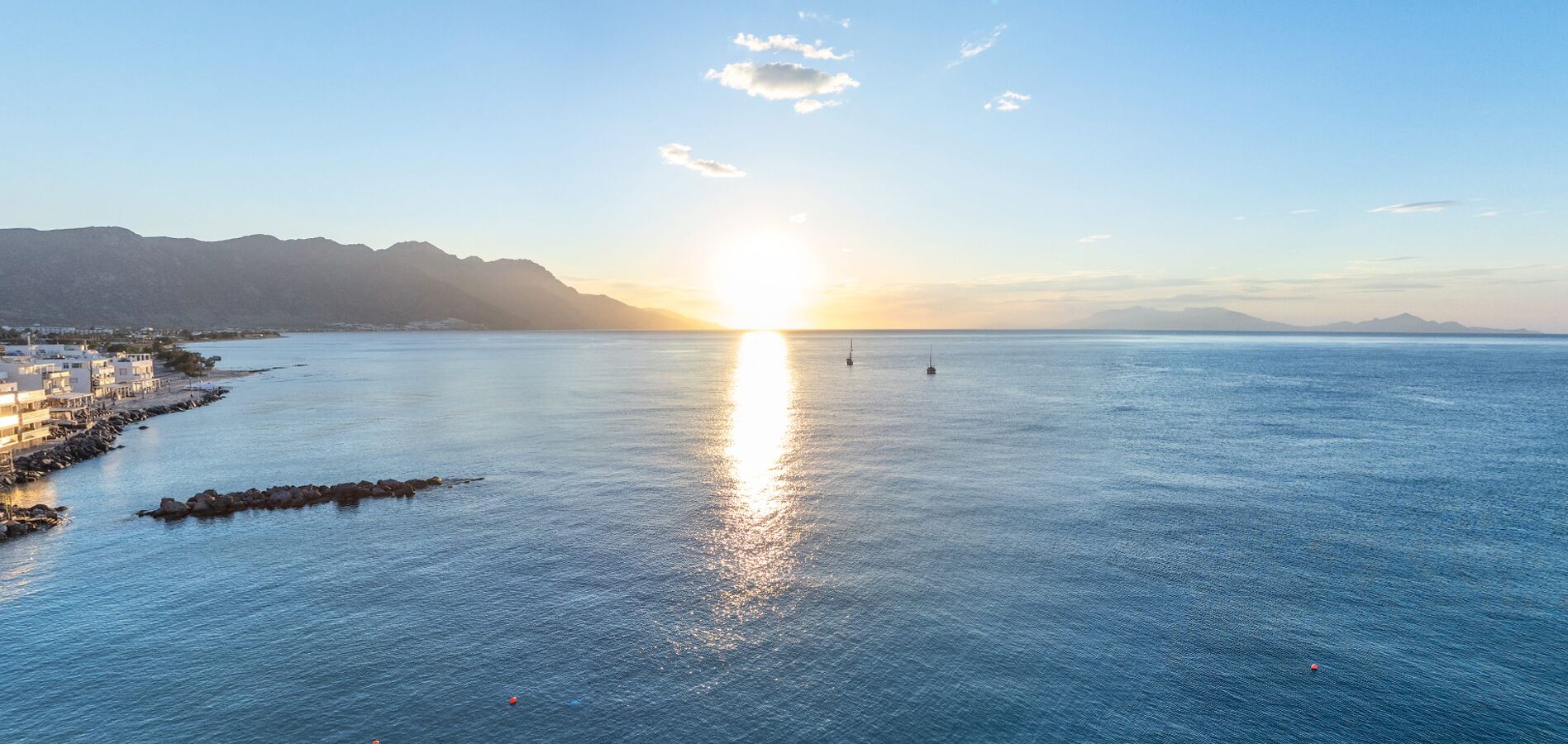Vista costera con amanecer sobre el mar, un rompeolas rocoso y una cadena montañosa distante.