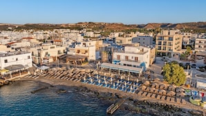 Un pueblo costero con una zona de playa que incluye tumbonas y sombrillas, edificios de techos planos, y un cielo azul despejado.