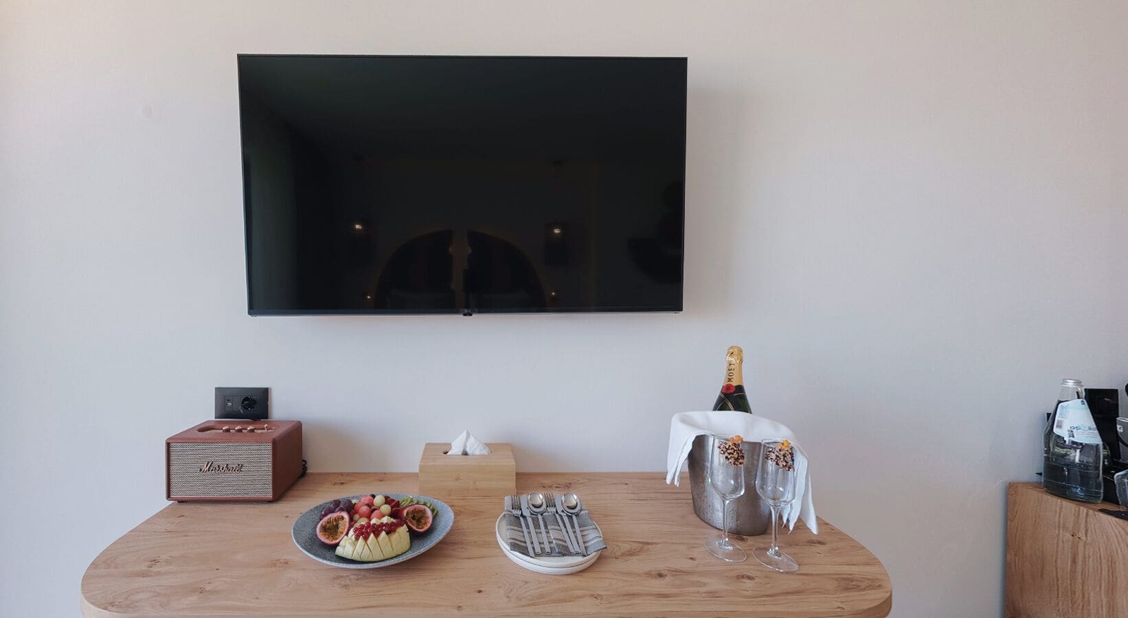 Una sala de estar moderna con una televisión de pantalla plana montada en la pared, una mesa de centro de madera con una bandeja de frutas, dos copas de champán y una radio antigua.