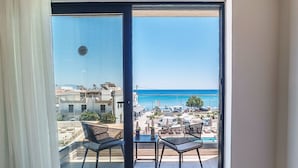 Un balcón con dos sillas y una mesa, con vistas a una playa y un puerto deportivo con barcos.