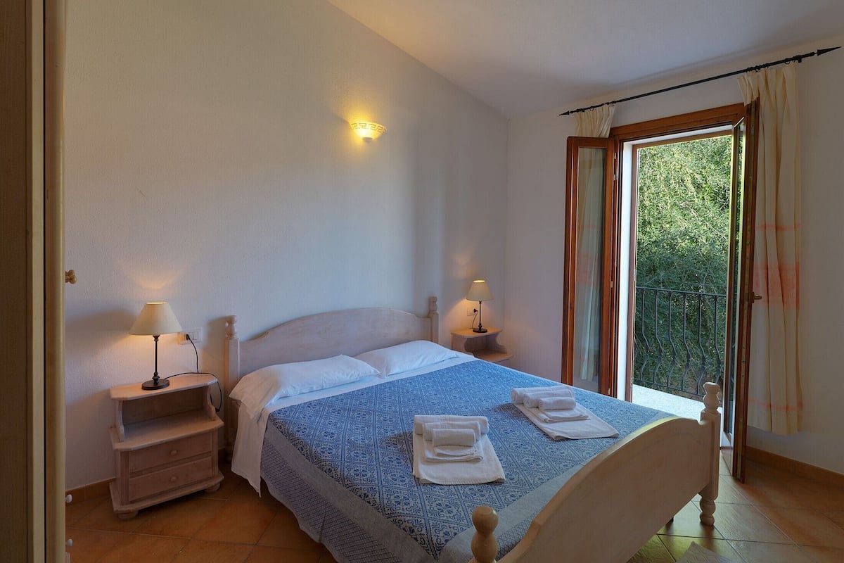 A bedroom with a bed, two bedside tables, a nightstand, a window with curtains, and a view of greenery outside.