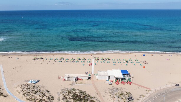 Une vue aérienne d’une plage avec des rangées de chaises longues et de parasols, une tour de surveillance et un petit bâtiment.
