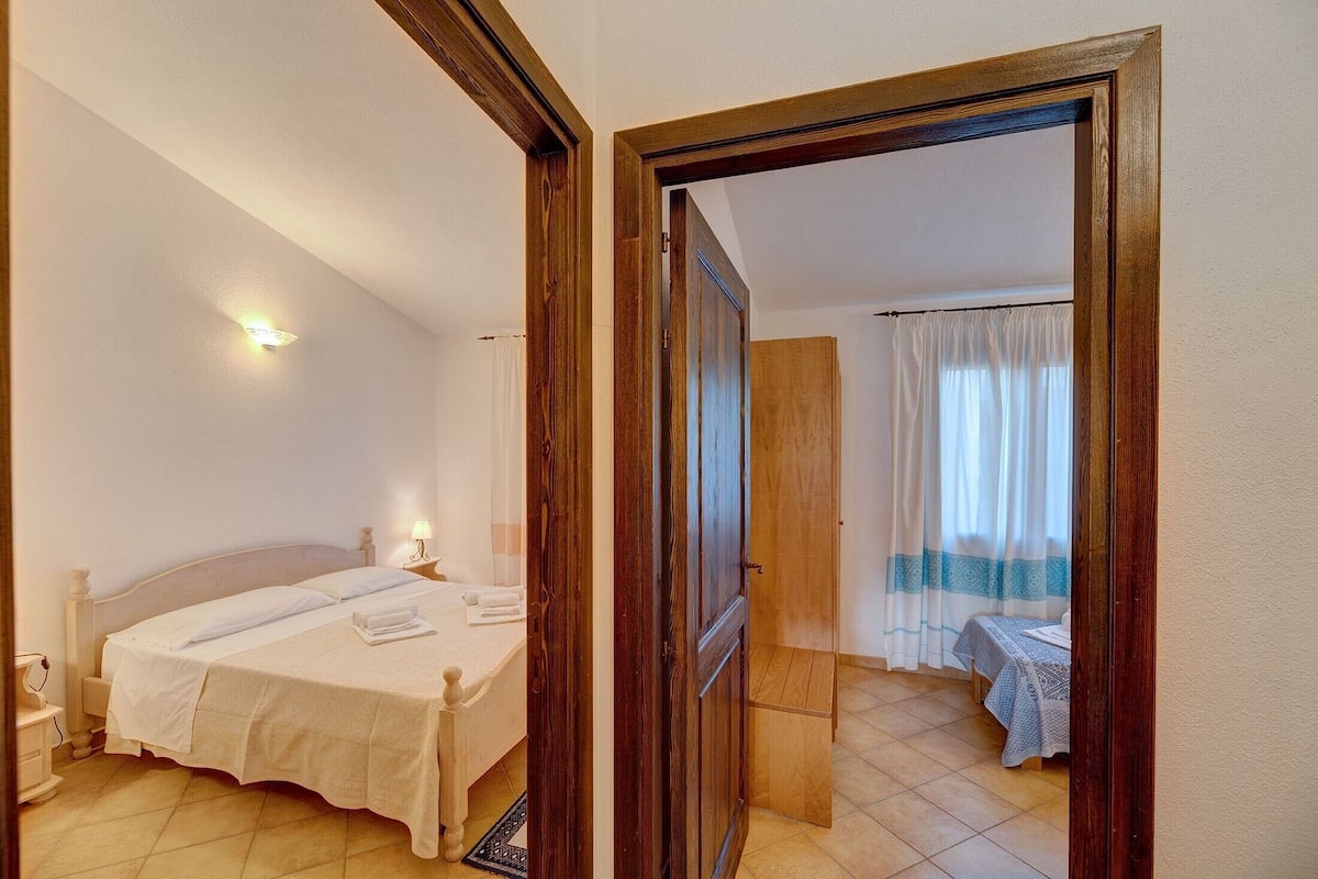 A bedroom with a bed, bedside table, and a door leading to another room.