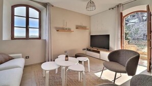 A modern living room with a sofa, a TV, a dining table, and a chair. There is a window with curtains, a hanging light fixture, and a view of the outside.