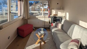 A living room with a sofa, a red ottoman, a glass coffee table, and a view of a snowy mountain landscape through large windows. - Attic apartment 2 minutes from the center, with a dream terrace (Leukerbad)
