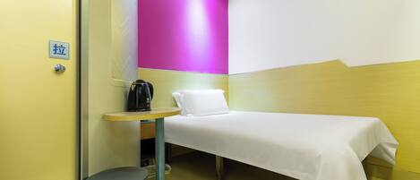 A small hotel room with a bed, a table with a kettle, a trash bin, and a wall with a bright pink accent.