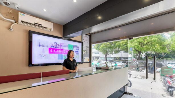 A reception desk with a digital display, a receptionist, and a view of parked cars and trees outside. - YIbaihotel (Shanghai)