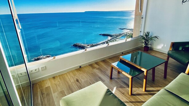 Un balcon avec vue sur la mer, une table en verre et des sièges confortables avec des coussins verts.