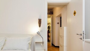 Ein Hotelzimmer mit einem Bett, einer Nachttischlampe, einem Wandregal, einer Tür mit einer Nummer und einem Flur, der in einen Küchenbereich führt.