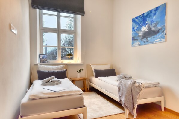 A bedroom with two beds, a window, a painting, and a bedside table.