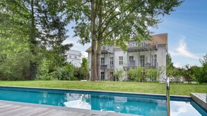 A swimming pool with a wooden deck, surrounded by greenery and a building with balconies.