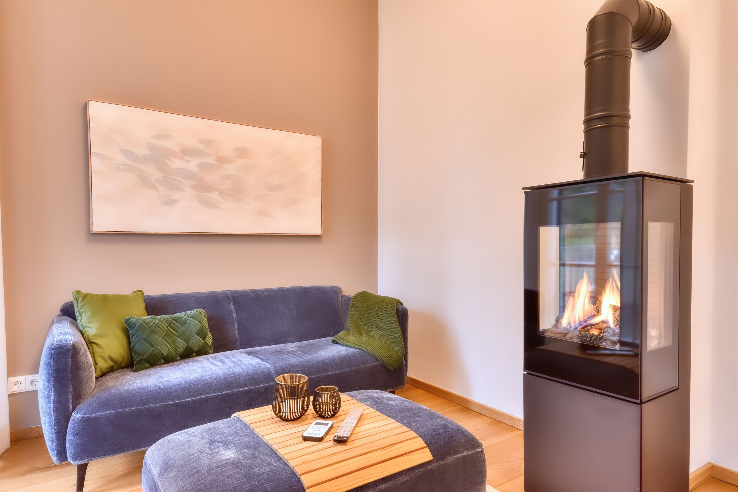 Ein modernes Wohnzimmer mit einem blauen Sofa, grünen Kissen, einem Couchtisch aus Holz mit zwei Kerzen und einem schwarzen Kaminofen.