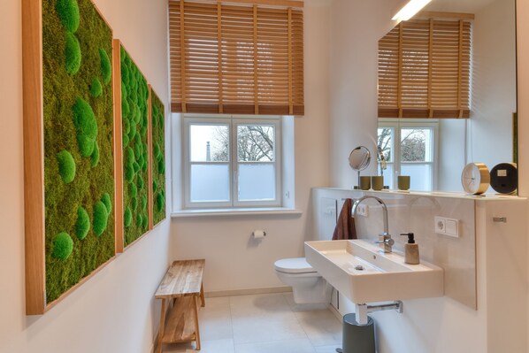 A modern bathroom with a white sink, mirror, and toilet. There is a wooden bench and a window with blinds.