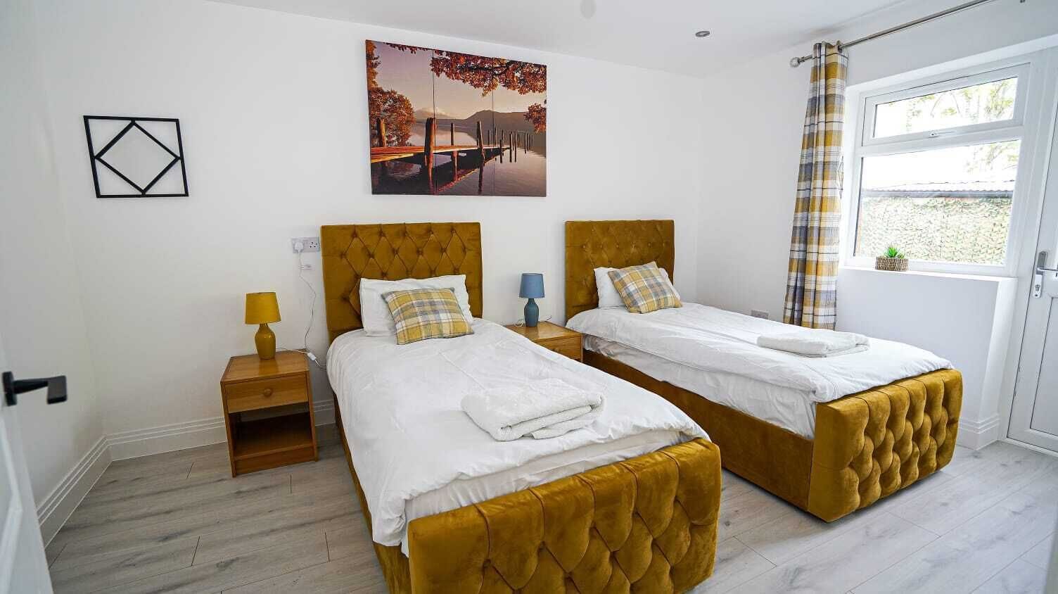 A double room with two beds, a painting, a window with curtains, and a wooden nightstand with a lamp.
