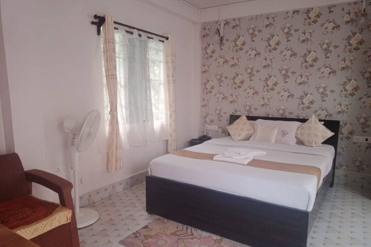A bedroom with a bed, a chair, a fan, and a window with curtains.