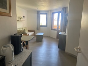 A compact hotel room with a bed, a small table, a kitchenette, and a window with curtains.