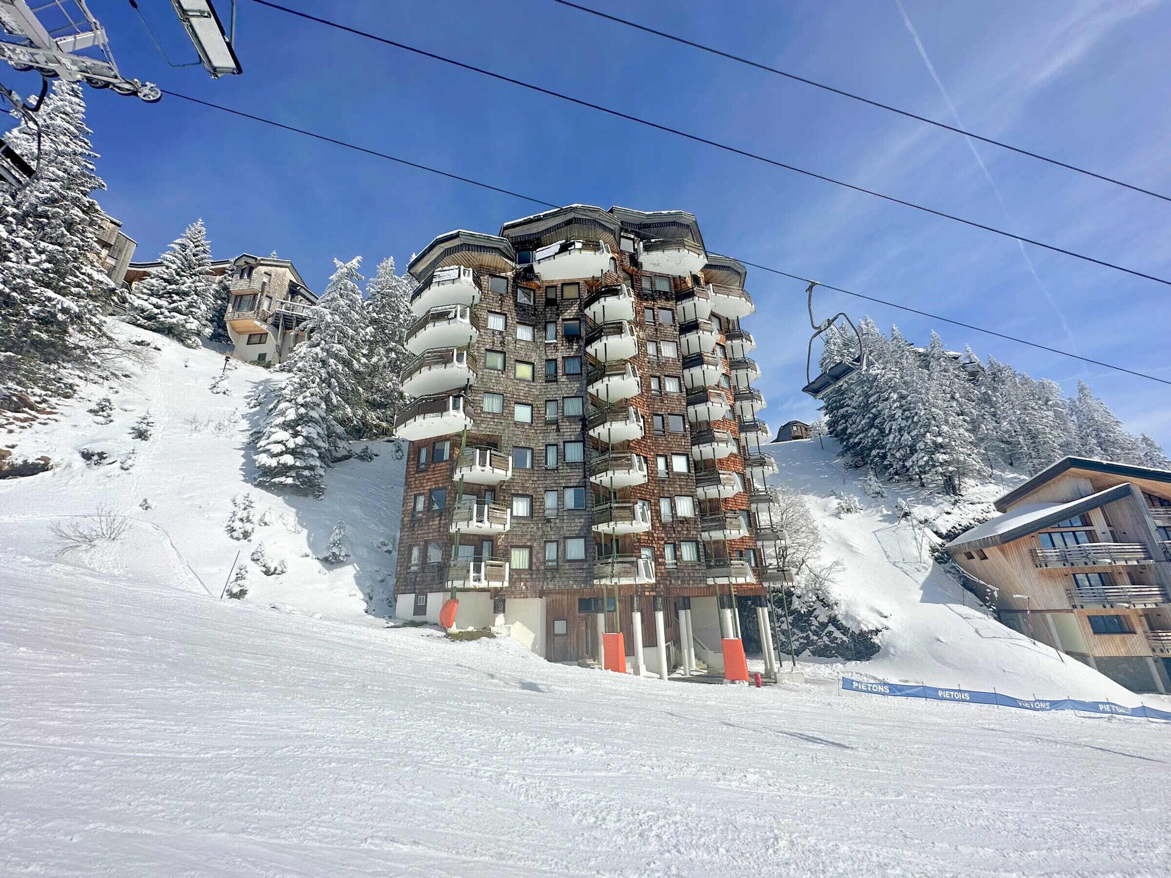 Un hotel de varios pisos con balcones, situado en una ladera nevada con telesillas al fondo.