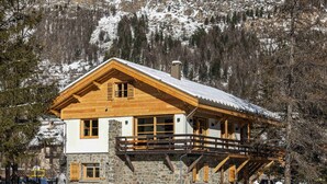 A chalet-style mountain lodge with a stone foundation, wooden exterior, and a balcony overlooking a snowy landscape.
