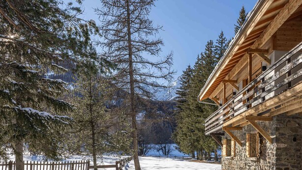 A snow-covered landscape with a wooden building featuring a stone foundation and a balcony.