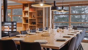 A dining area with a long wooden table set for a meal, surrounded by chairs, with a fireplace and bookshelves in the background.