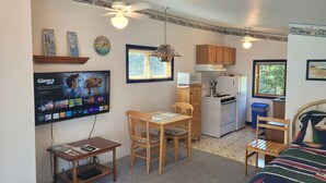 A compact kitchen with a microwave, stove, and refrigerator. There is a dining table with chairs, a small TV mounted on the wall, and a ceiling fan.