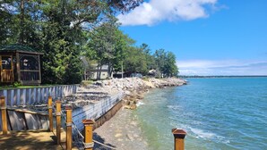 A wooden boardwalk leading to a rocky shoreline with a clear view of the water and a building with a green roof in the background.