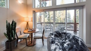 A cozy room with a round table, two chairs, a potted plant, a fluffy rug, and a large window overlooking a deck.