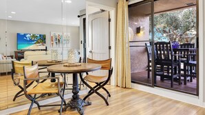 A dining area with a round table and chairs, a television, and a view of an outdoor patio with chairs.