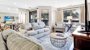 A modern living room with a sofa, armchairs, a coffee table, and a TV. There is a dining area with a table and chairs, and a large window providing a view of the outdoors.