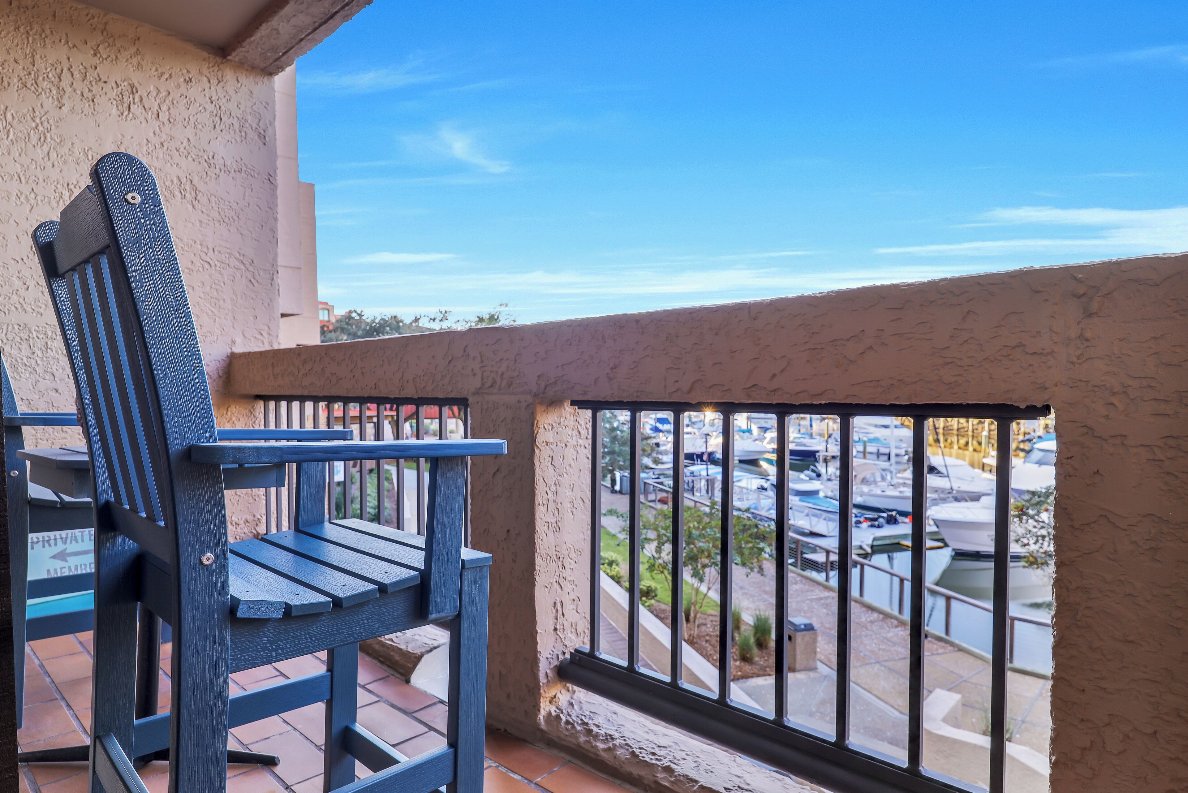 A balcony with a blue chair, a view of a marina with boats, and a clear blue sky.