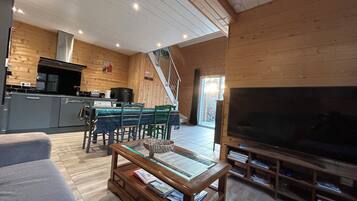 Un salon moderne avec un décor intérieur en bois, un coin repas et une cuisine équipée d’une grande télévision et de bibliothèques.
