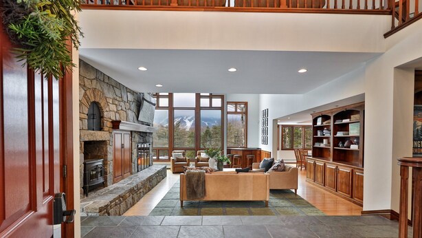 A spacious living area with a stone fireplace, large windows offering mountain views, a cozy seating area, and a wooden bookcase.