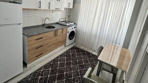 A compact kitchen with white cabinets, a gas stove, a sink, and a washing machine. There's a dining area with a table and chairs. - AVRUPA Suit - otel (Bandirma)