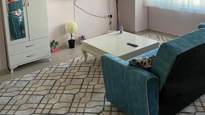A room with a teal sofa, a white TV mounted on the wall, a small table, a cabinet with a mirror, and a window with sheer curtains. - AVRUPA Suit - otel (Bandirma)