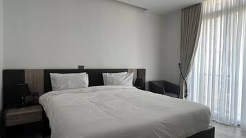 A modern hotel room with a large bed, two bedside tables, a nightstand, and a window with sheer curtains.