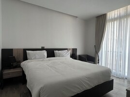 Una habitación de hotel moderna con una cama grande, dos mesitas de noche y un ventanal con cortinas translúcidas.