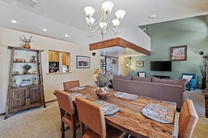 A dining area with a wooden table and chairs, a chandelier, and a kitchen in the background.