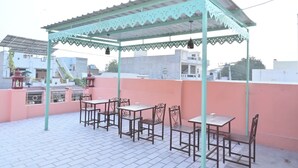 A rooftop patio with a green pergola, several tables and chairs, and a view of surrounding buildings.