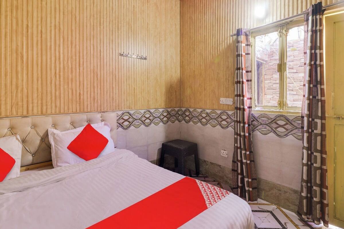 A bedroom with a bed, pillows, a small stool, a window with curtains, and a patterned wall.