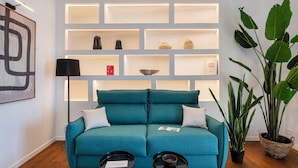 Een smaragdgroene bank met twee kussens, een zwarte vloerlamp en een boekenkast met diverse spullen.