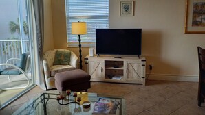 A living room with a glass coffee table, a sofa, a television on a wooden stand, a lamp, and a window with blinds.