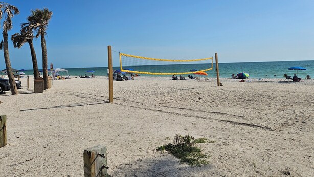 A beach volleyball net on a sandy beach with palm trees and umbrellas.