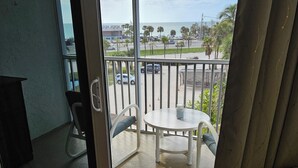 A balcony with a view of the ocean and parked cars, a white table with chairs, and a patterned sofa.