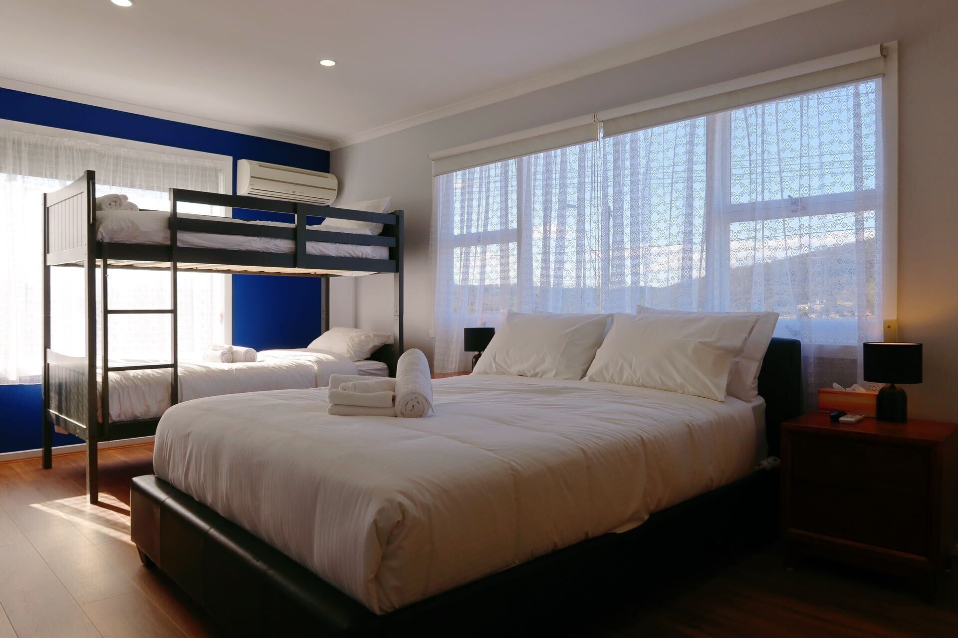 A bedroom with bunk beds, a large window with patterned curtains, a bedside table with a lamp, and a wooden floor.