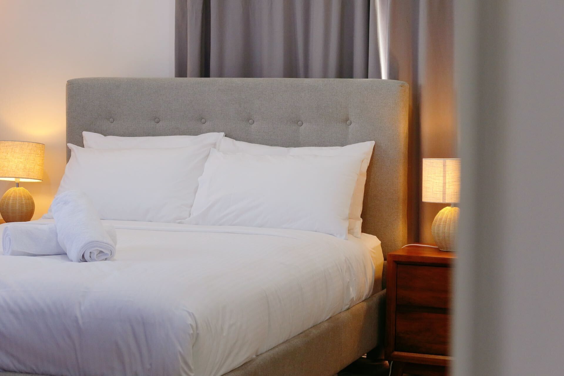 Una cama impecable con sábanas y almohadas blancas, un cabecero tapizado en gris y dos lámparas de noche sobre mesitas de madera.