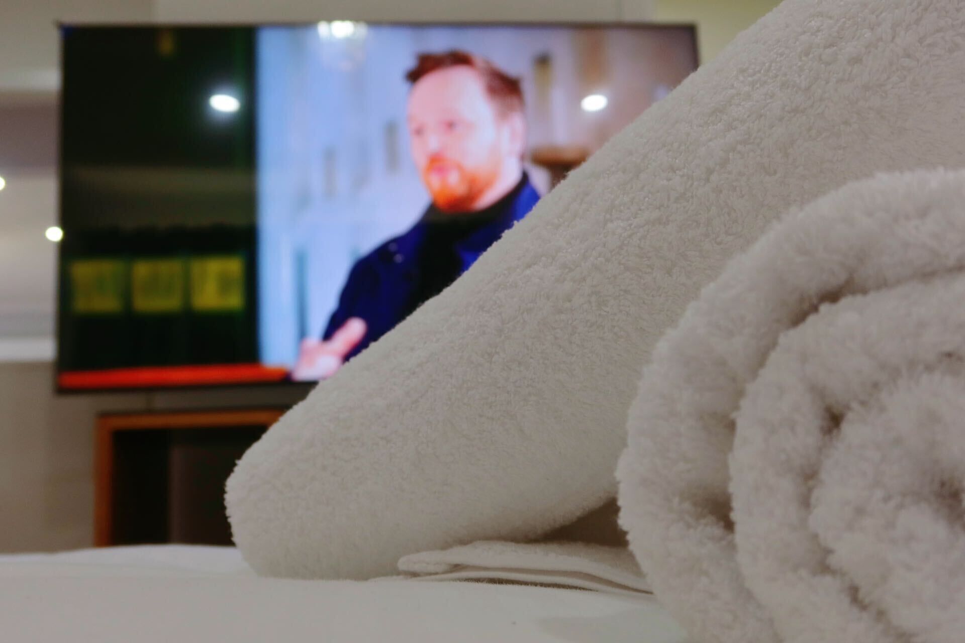 Habitación con televisor mostrando a una persona, una toalla blanca enrollada y una cama.