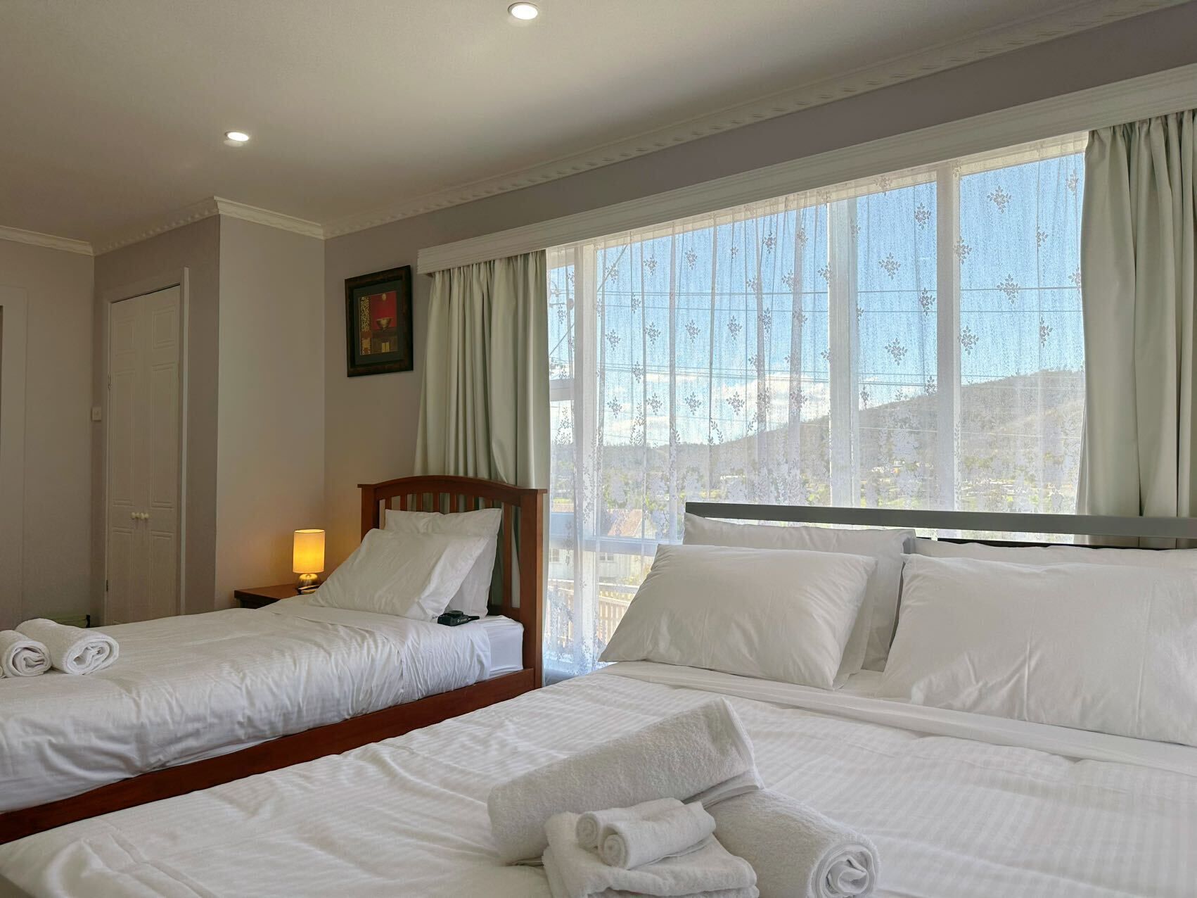 Habitación de hotel con dos camas, una mesita de noche con lámpara, un cuadro en la pared y ventanas amplias con cortinas.