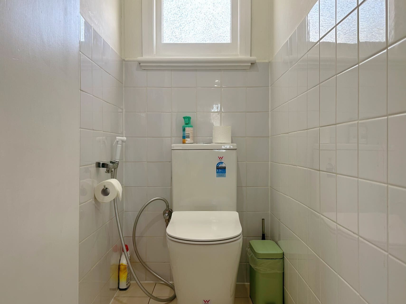 Un baño pequeño y limpio con baldosas blancas, un inodoro, un cesto de basura y una ventana sobre el inodoro.