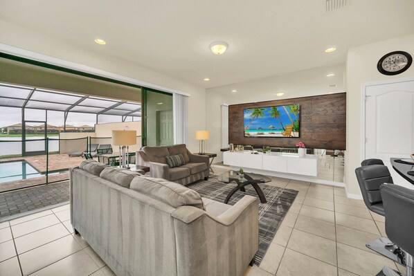 A modern living room with a large sofa, a coffee table, and a TV mounted on a wooden wall. There is a pool visible through large windows.
